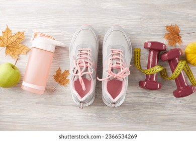 Autumn muscle-building routine idea. Top view shot of trendy shoes, tape measure, dumbbells, fresh apple, water bottle, pumpkin, autumn leaves on light wooden background - Shutterstock ID 2366534269