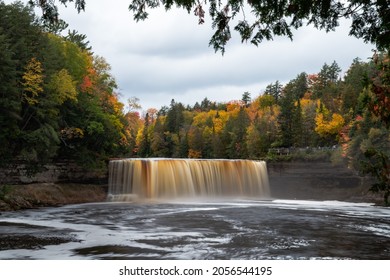 Autumn photograph of Upper Tahquamenon Falls waterfall in Michigan with water cascading into the river below surrounded by evergreen trees and fall colored foliage with cloudy sky above.