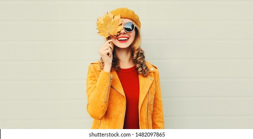 Autumn portrait of beautiful happy smiling young woman with yellow maple leaves wearing a french beret hat, sunglasses and jacket on gray background Stock Photo