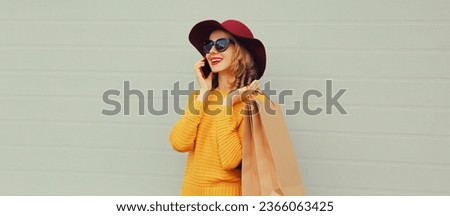 Autumn portrait of beautiful happy smiling young woman with shopping bag calling on mobile phone looking away wearing hat, yellow sweater on gray background