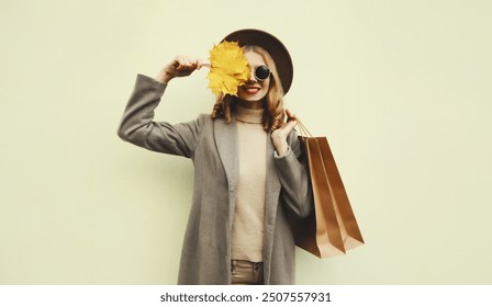 Autumn portrait of beautiful happy young woman with shopping bag, stylish girl holding yellow maple leaves in round hat, coat on brown studio background - Powered by Shutterstock