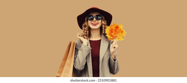 Autumn portrait of beautiful happy young woman with shopping bag, stylish girl holding yellow maple leaves in round hat, coat on brown studio background - Powered by Shutterstock