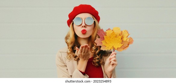 Autumn portrait of beautiful young woman model with yellow maple leaves blowing her lips with lipstick wearing a red french beret on gray background