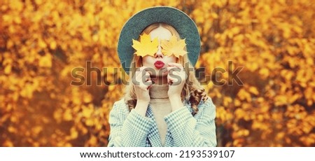 Autumn portrait of beautiful young woman covering her eyes with yellow maple leaves blowing her lips wearing round hat in the park