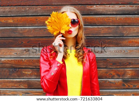 Autumn portrait of beautiful young woman with yellow maple leaves blowing her lips with lipstick wearing red heart shaped sunglasses on wooden wall background