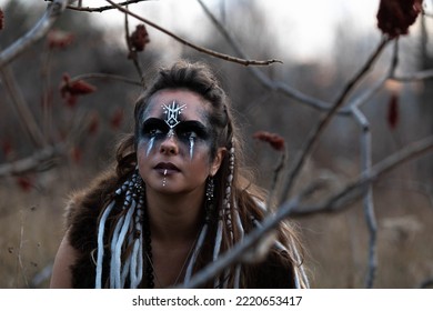 Autumn Portrait Of A Female Viking Squatting In A Forest. She Is Wearing Face Paint, Leather And Fur. She Has Her Hair In Braids. 