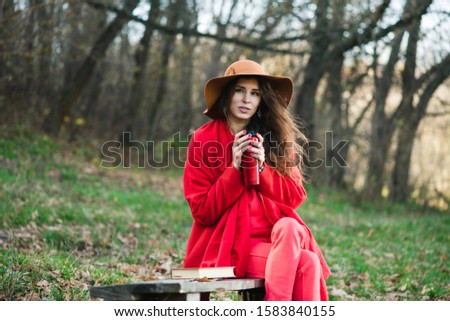 Autumn portrait of girl hold red thermos. Beautiful young brunette wears a red coat, scarf and brown wide-brimmed hat.