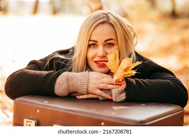 Autumn Portrait Of Happy, Beautiful 35 - Year - Old Blonde Woman In Black Coat And With Yellow Leaves In Her Hands. She Holds Her Head In Her Hands, Lying On Suitcase In Autumn Park.