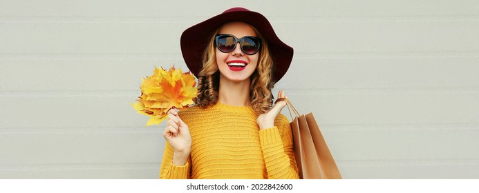Herbstporträt einer glücklich lächelnden jungen Frau mit Einkaufstaschen und gelben Ahornblättern mit Pullover und Hut auf grauem Hintergrund