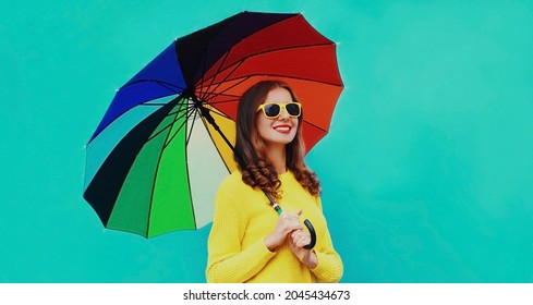 Herbstporträt einer glücklich lächelnden jungen Frau, die einen bunten Schirm mit einem gelben Strickpullover auf blauem Hintergrund hält