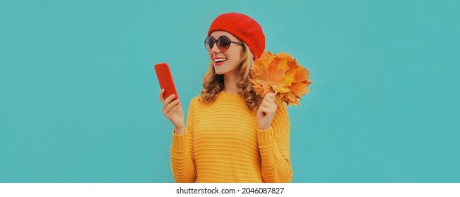 Autumn Portrait Of Happy Smiling Young Woman With Smartphone And Yellow Maple Leaves Wearing French Red Beret On Blue Background