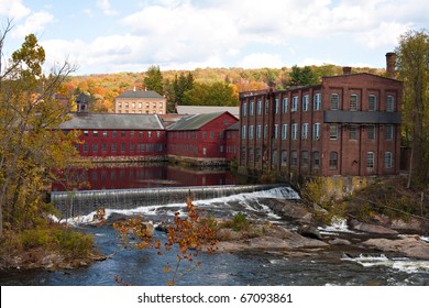 An Autumn river scene with some falls and old mill buildings alongside with colorful foliage on the horizon.