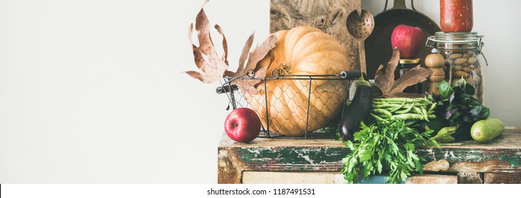 Autumn seasonal food ingredients, kitchen utensils. Vegetables, pumpkin, apples, canned food, fallen leaves over rustic chest of cupboard, copy space, wide composition. Thanksgiving dinner preparation