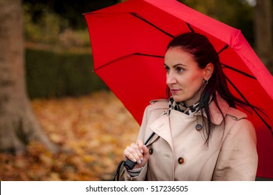 Autumn Setting With A Mature Woman Standing Outdoors In A Park Next To A Tree In The Fall, Wearing A Trench Coat Holding And A Red Umbrella Not Looking At The Camera, Admiring The Fall Foliage