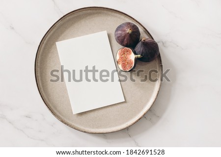 Autumn still life. Blank greeting card mockup, cut figs fruit and ceramic plate on white marble table background. Fall and Thanksgiving concept, styled stock flat lay photography. Top view.