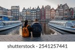 As the autumn sun sets, a couple relaxes by the serene canal in Amsterdam, surrounded by iconic colorful buildings and peaceful boats. The gentle fall breeze enhances their intimate moment.