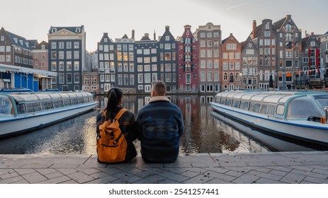 As the autumn sun sets, a couple relaxes by the serene canal in Amsterdam, surrounded by iconic colorful buildings and peaceful boats. The gentle fall breeze enhances their intimate moment. - Powered by Shutterstock