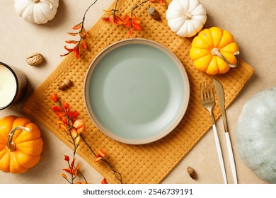 Autumn table setting with empty plate, pumpkins, leaves, acorns, and cutlery on a beige background. Perfect for Thanksgiving holiday celebration.