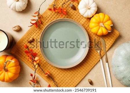 Autumn table setting with empty plate, pumpkins, leaves, acorns, and cutlery on a beige background. Perfect for Thanksgiving holiday celebration.