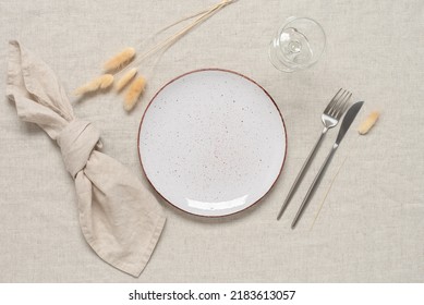 Autumn table setting. A white plate, cutlery and dry grass lagurus on a beige linen textile background. Top view, flat lay. Selective focus.