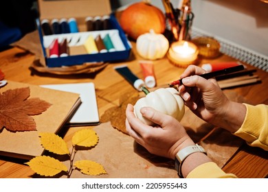 Autumn Thanksgiving Pumpkins Craft. DIY Fall Crafts For Adults. Female Hands Draw Patterns On White Pumpkin Against Autumn Decor Background On The Table At Home