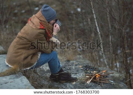 Autumn. Tourist girl outdoors in the nature with a backpack and a thermos for the trip travel. A woman with a bag and a hat in close-up is resting by the fire with hot tea. Warm outerwear.