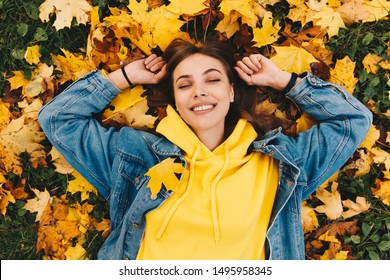 Autumn walk. Woman portrait. Happy girl in yellow hoodie and jean jacket is smiling while lying with her eyes closed on the ground in the park; top view