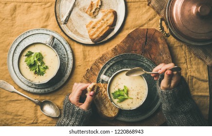 Autumn, Winter Home Dinner. Flat-lay Of Fall Warming Celery Cream Soup And Female Hands In Sweater With Spoon Over Linen Tablecloth, Top View. Comfort Food, Vegetarian, Healthy And Slow Food Concept