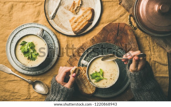 Autumn, Winter home dinner. Flat-lay of Fall\
warming celery cream soup and female hands in sweater with spoon\
over linen tablecloth, top view. Comfort food, vegetarian, healthy\
and slow food concept