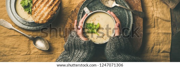 Autumn, Winter home dinner. Flat-lay of Fall\
warming celery cream soup, grilled bread and female hands over\
linen tablecloth, top view, wide composition. Comfort food, healthy\
and slow food concept