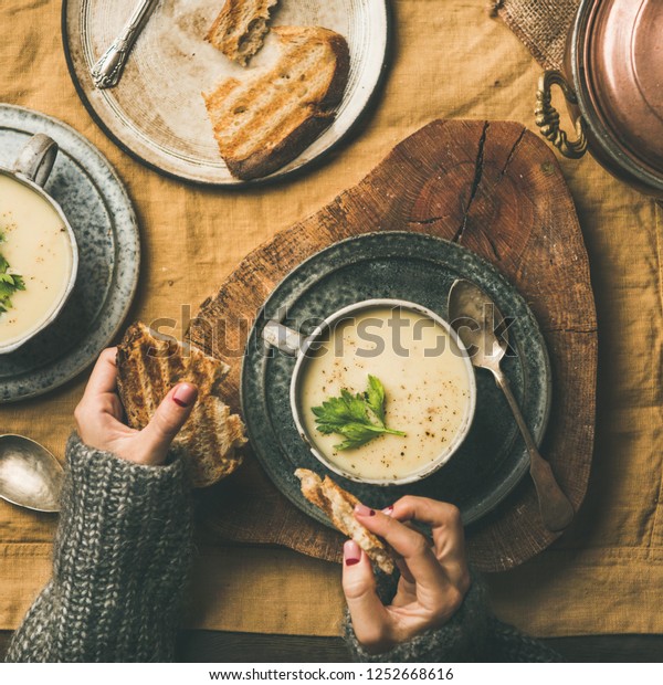 Autumn, Winter home dinner. Flat-lay of Fall\
warming celery cream soup and female hands with bread over linen\
tablecloth, top view, square crop. Comfort food, vegetarian,\
healthy and slow food\
concept