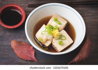 Autumn And Winter Hot Japanese Recipe. Fresh Hot Tofu Served With Daikon, Green Onion,  Chive Ginger And Soy Sauce Topping  On Ceramic Traditio Blow  With Chopsticks And Autumn Leaves, Wooden Table