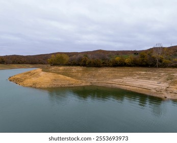 Autumnal Landscape of a Dried-out Lakeside. High-angle view of a serene lake shoreline receding with exposed, light brown earth, bordered by a hillside of autumn foliage.  - Powered by Shutterstock