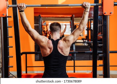 Average Plan, Back View. A Muscular Man Dressed In Black Sportswear Is Training Muscles, Biceps And Triceps - With An Effort He Lifting The Bar. Fitness And Sport. Healthy Lifestyle Concept