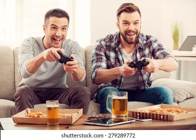 The Avid Gamers. Two Young Happy Men Playing Video Games While Sitting On Sofa