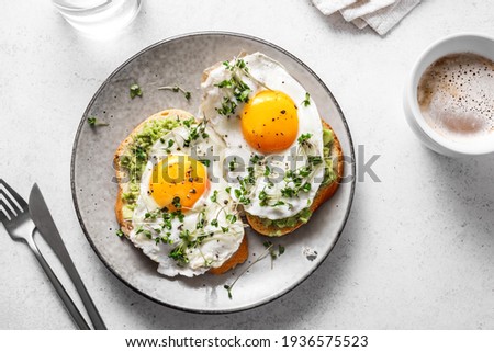 Avocado Egg Sandwiches and coffee for healthy breakfast. Whole grain toasts with mashed avocado, fried eggs and organic microgreens on white table.