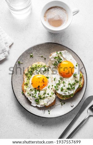 Avocado Egg Sandwiches and coffee for healthy breakfast. Whole grain toasts with mashed avocado, fried eggs and organic microgreens on white table.