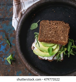 Avocado and ricotta rye sandwich with fresh herds on black clay tray with checkered kitchen towel over dark wooden textured background. Healthy eating theme. Top view. Square image