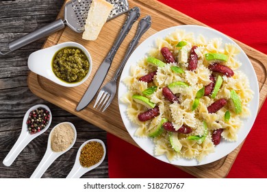 Avocado, Sausages Bow Tie Pasta Warm Salad With Basil Sprinkled With Grated Parmesan Cheese On White Plate On Chopping Board. Spices  And Grater On Dark Wooden Background, Close-up, View From Above