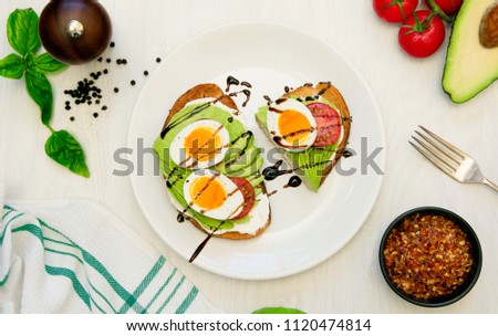 Avocado and soft boiled egg on whole grain toast. On white plat with fork and tea bowl in background. pepper, basil and salt in one corner. Half an avocado and vine tomatoes. 