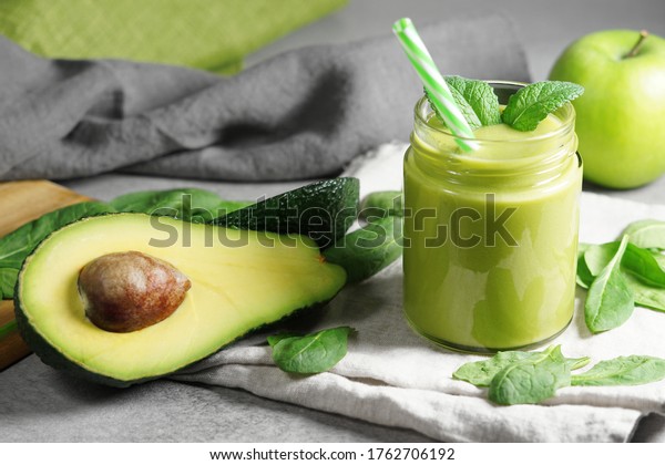 Avocado spinach and green apple smoothie healthy\
and refreshing tropical drink close-up view of a glass jar with\
mint leaves and sliced avocado fruit, apple and spinach leaves on\
grey kitchen board.