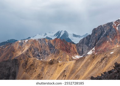 Awesome aerial view to sunlit sharp rocks on ridge of vivid colors and snowy peak in low clouds. Scenic misty landscape with colorful sheer crags against snow-capped mountain top under gray rainy sky. - Powered by Shutterstock