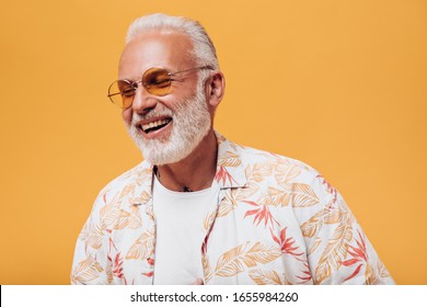 Awesome man in orange eyeglasses laughing on isolated background. Gray-haired adult with beard in light outfit is smiling into camera
