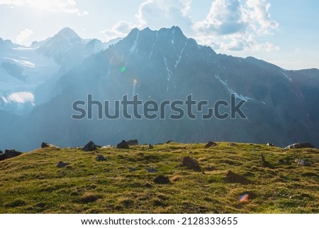 Awesome mountain scenery with big sharp stones in green grass near abyss edge with view to giant mountain in sunlight. Scenic sunny alpine landscape with sharp rocks in grasses near edge of precipice.