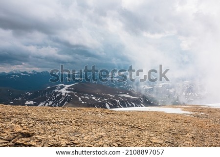 Awesome top view through clouds to high snowy mountains. Scenic landscape with beautiful snow mountains in low clouds. Atmospheric alpine view from stone hill to snow mountain range with low clouds.