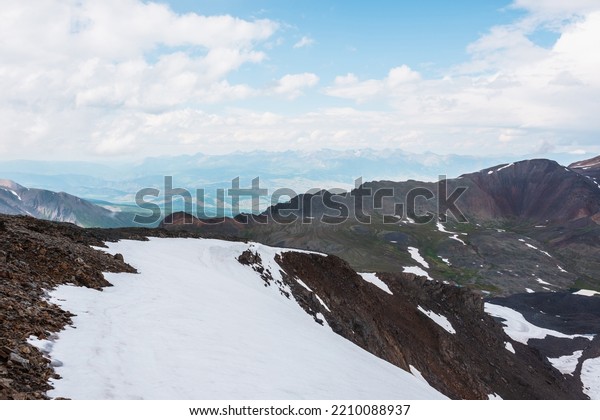 Awesome view from sharp rocks with snow cornice\
to multicolor mountain valley. Top view from stone hill with snow\
to high mountains under cloudy sky. Beautiful mountain landscape at\
very high altitude