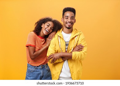 Awesome when boyfriend is best friend. Portrait of charming friendly african american woman leaning on guy touching his shoulder feeling happy they together and she can rely on posing orange wall