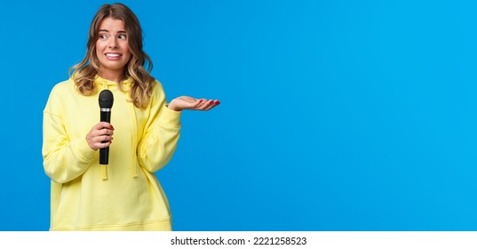 Awkward Cute Blond Girl Dont Know Lyrics As Singing Karaoke, Looking At Friend Nervous Smile, Embarrassed Sing In Fron Of People, Hold Microphone, Standing Blue Background.