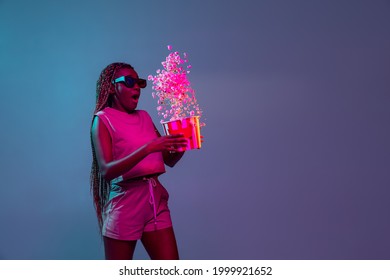 Awkward situation. . Young pretty african woman in 3d glasses throwing popcorn from bucket isolated on blue studio background in neon light. Copyspace for ad.