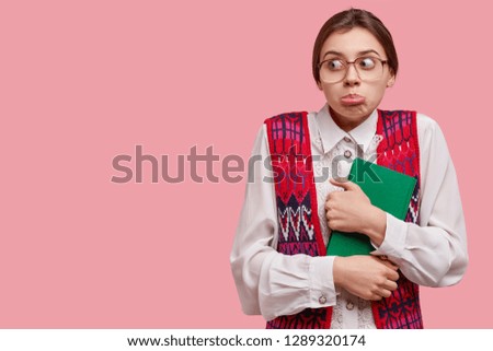 Awkward woman in funny eyeglasses and old clothes looks suspiciously aside, purses lower lip, carries notebook, notices something surprising on blank space, models in studio against pink background